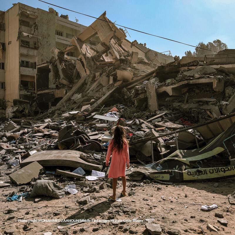 In Gaza, a girl stands in front of a pile of rubble and destroyed buildings. 